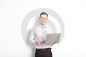 Smiling confident young man, no tie, holding grey laptop device and typing while standing against solid white wall. Wireless inter