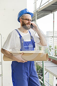 Smiling construction worker in work outfit and helmet on head talking on the phone. Work at high altitude. Scaffolding in the back