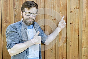 A smiling, contented man shows with one hand on a wooden, painted old wall, and the other makes a gesture, a thumb up. Isolated