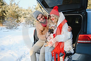 Smiling couple with dog sitting in open SUV car trunk in snowy forest. Enjoying each other in active winter holidays