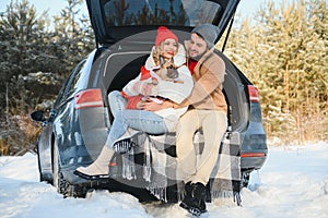 Smiling couple with dog sitting in open SUV car trunk in snowy forest. Enjoying each other in active winter holidays