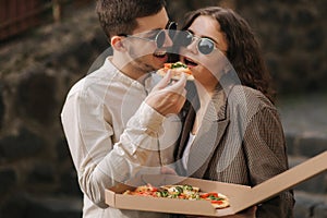 Smiling couple eating pizza outdoors. Man hol pizza box and take slice of vegan pizza. Stylish couple takeway pizza
