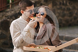 Smiling couple eating pizza outdoors. Man hol pizza box and take slice of vegan pizza. Stylish couple takeway pizza