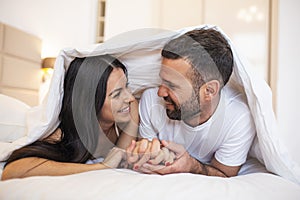 Smiling couple going to kiss under sleeping sheets in the morning. In love young happy couple lying in bed in the morning