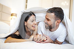 Smiling couple going to kiss under sleeping sheets in the morning. In love young happy couple lying in bed in the morning