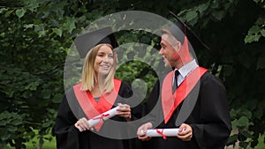 Smiling couple of graduates walking in park, holding diplomas and talking