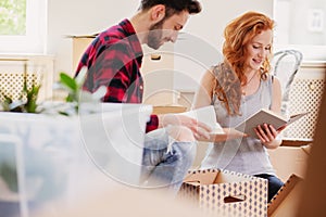 Smiling couple reading books while unpacking stuff after relocation to new flat