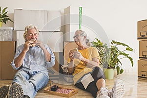 Smiling couple of senior people white hair working in the new empty apartment with moving boxes, sitting on the floor having break