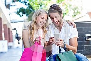 Smiling couple with shopping bags sitting and using smartphone