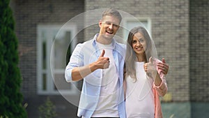 Smiling couple showing thumbs up, standing against house, real estate purchase