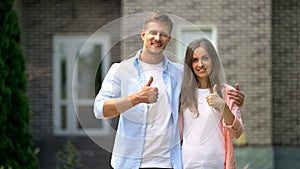 Smiling couple showing thumbs up, standing against house, real estate purchase