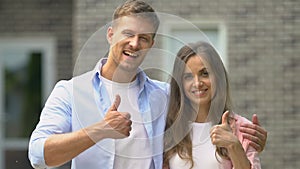 Smiling couple showing thumbs up standing outside house, real estate agency