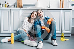 smiling couple sitting on floor and leaning on kitchen counter after cleaning
