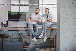 smiling couple sitting on kitchen counter with cups of coffee in morning