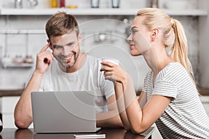 smiling couple sitting in kitchen with laptop and cup