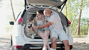 Smiling couple sitting in the open trunk car and petting Akita Inu