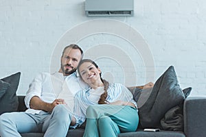 smiling couple sitting on sofa during the summer heat at home with air conditioner