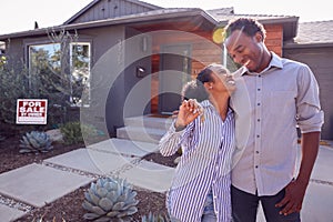 Smiling Couple Standing Outdoors In Front Of House With For Sale Sign In Garden Holding Keys