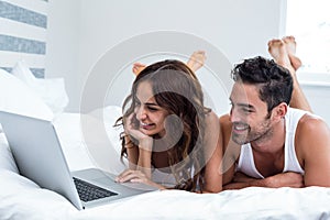 Smiling couple using laptop while lying under blanket