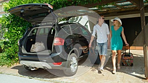 Smiling couple walks out of their house to pack their bags in the black car.