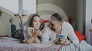 Smiling couple watching TV while lying in bed and drinking coffee at home in the morning