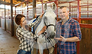 Smiling couple with white horse standing at stabling indoor