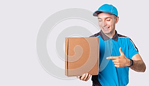 Smiling courier in blue uniform and a cap is pointing to the parcel he must deliver it to destination.