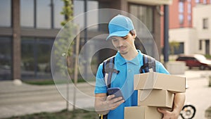 Smiling Courier Going Along Building Holding the Paper Boxes and Mobile Phone in Hand. Delivery Man Looking for Delivery