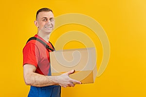 Smiling courier man in blue jumpsuit and red T-shirt holding craft paper box. Isolated on yellow background. Copy space, mock up