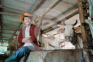 smiling cowboy man wearing a cowboy hat while sitting near the cow shed with crossed hands