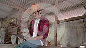 smiling cowboy man wearing a cowboy hat while sitting near the cow shed with crossed hands