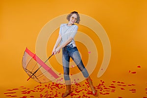 Smiling curly girl with umbrella posing on yellow background. Slim young lady in denim pants standing on the floor