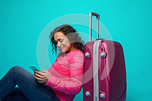 Smiling curly-haired beautiful woman in pink jacket using phone, browsing websites, booking tickets, making online reservation