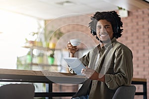 Smiling curly young black guy in wireless headphones holds cup of tea, listens music, enjoy free time
