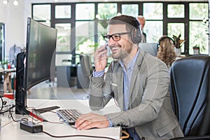 Smiling customer support operator wearing headset talking with client while working on computer in office.