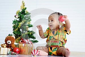 Smiling cute African baby kid in colorful dress sits near Christmas tree and gift box present in white wall living room, little