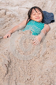 Smiling cute Asian kid, child, little girl lay down on the sand beach in sunny day