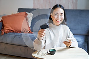 Smiling cute asian woman using credit card and smartphone, paying bills online, holding mobile phone, looking at camera