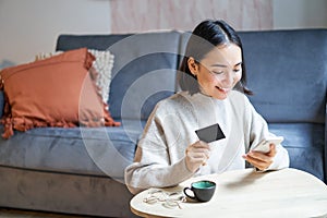 Smiling cute asian woman using credit card and smartphone, paying bills online, holding mobile phone, looking at camera