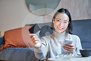 Smiling cute asian woman using credit card and smartphone, paying bills online, holding mobile phone, looking at camera