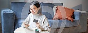 Smiling cute asian woman using credit card and smartphone, paying bills online, holding mobile phone, looking at camera