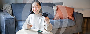 Smiling cute asian woman using credit card and smartphone, paying bills online, holding mobile phone, looking at camera