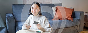 Smiling cute asian woman using credit card and smartphone, paying bills online, holding mobile phone, looking at camera