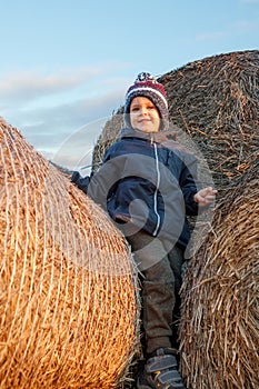 A smiling cute boy with a knitted hat stands on hay bales in the evening sunlight