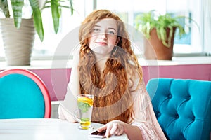 Smiling cute girl is resting in cafe, holding smartphone in hand. Woman with long red curly hair