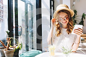 Smiling cute girl in straw hat relaxing in cafe beside glass door holding smartphone in hand. Indoor portrait of pretty