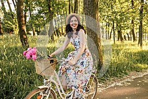 Smiling cute happy girl in dress and hat riding retro bicycle with flowers in the buncket. Green sunny park is around