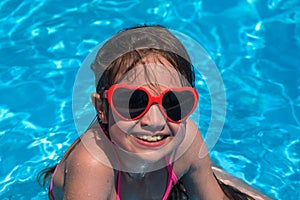 Smiling cute kid girl in heart shaped sunglasses in swimming pool on sunny day