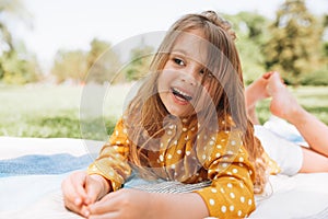 Smiling cute little girl lying on the blanket at green grass, talking with her parents. Adorable child having fun outdoors during