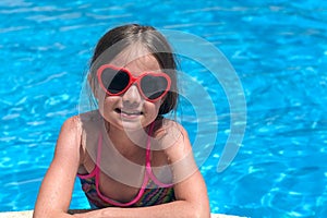 Smiling cute little girl in sunglasses in swimming pool on sunny day. Childhood and summer